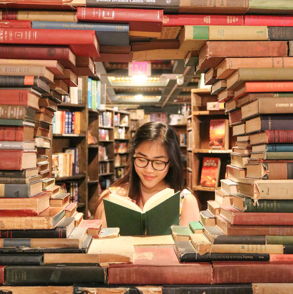 junge Frau liest Buch und schaut durch einen großen Berg Bücher